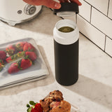 Person putting black silicone leakproof lid on Porter Insulated Charcoal Waterbottle with white top, next to kitchen counter and strawberries. 