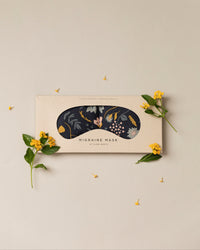 Migraine Mask by Slow North. Strap free cotton weighted eye pillow to be used hot or cold to sooth headaches and tired eyes. Navy and floral print. Inside box surrounded by yellow flowers.