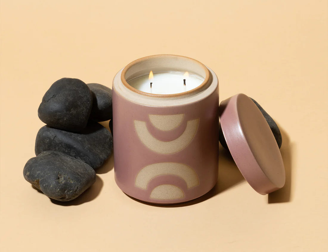Paddywax blush colored Mandarin Mango Candle. Lit candle with lid off and surrounded by black rocks.