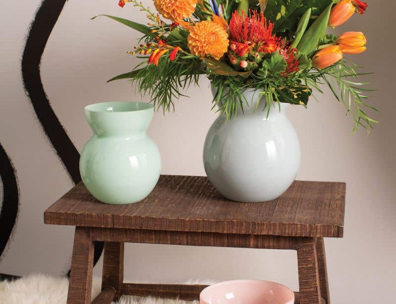 Glenna Vases handcrafted with powdery glaze. Three shown in light green, light pink, and light blue holding an orange bouquet. Set in home on wooden stool and fur rug. 