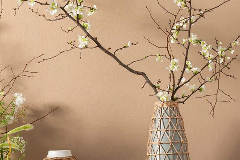 Three Lissome Collection of speckled neutral toned earthenware vases with woven rattan accents. Holding woody plant with white flowers and sitting on side table. 