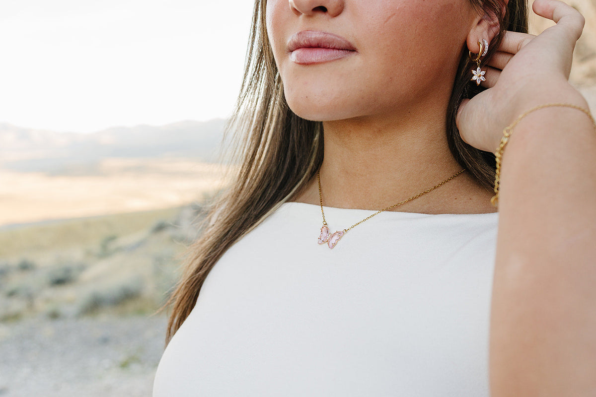 Woman in a white top wearing a gold butterfly necklace with a pink crystal butterfly, a gold flower drop earring and a gold chain bracelet, with the desert behind her