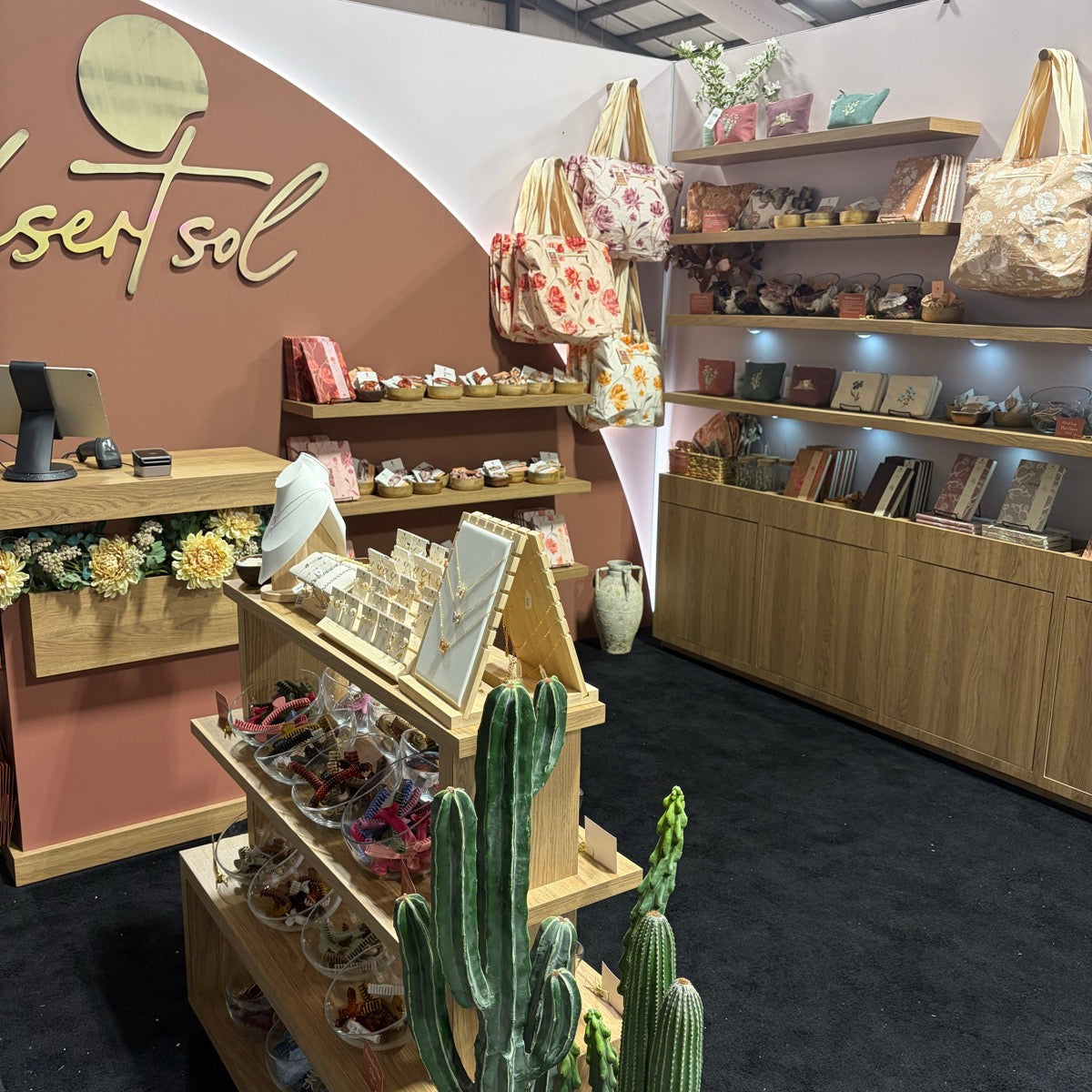 The Desert Sol booth at the Pinners Conference in Colorado, a curved terracotta wall with the desert sol name in gold script, light oak shelves of printed totes, journals and hair clips, and tall faux cactus by the jewelry stand