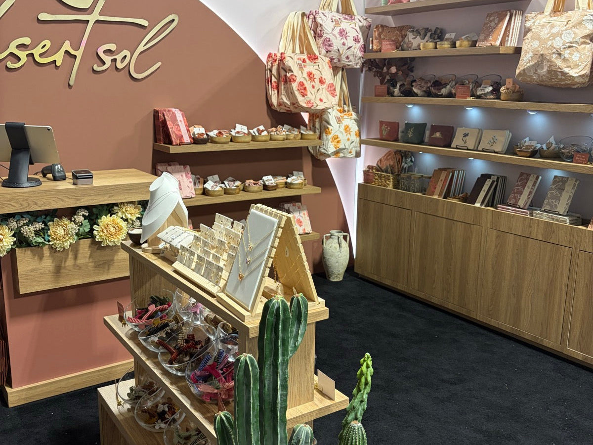 The Desert Sol booth at the Pinners Conference in Colorado, a curved terracotta wall with the desert sol name in gold script, light oak shelves of printed totes, journals and hair clips, and tall faux cactus by the jewelry stand