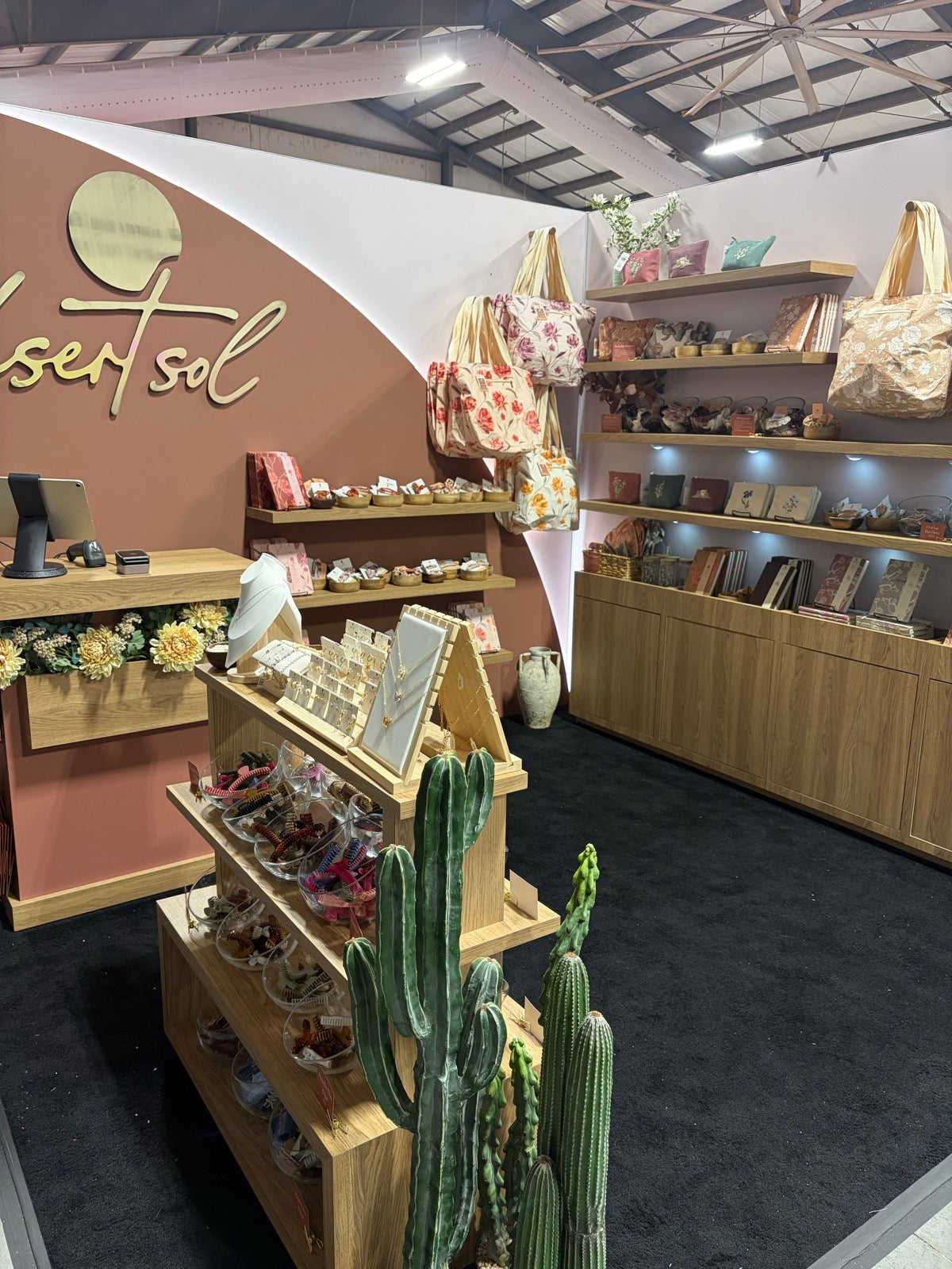The Desert Sol booth at the Pinners Conference in Colorado, a curved terracotta wall with the desert sol name in gold script, light oak shelves of printed totes, journals and hair clips, and tall faux cactus by the jewelry stand