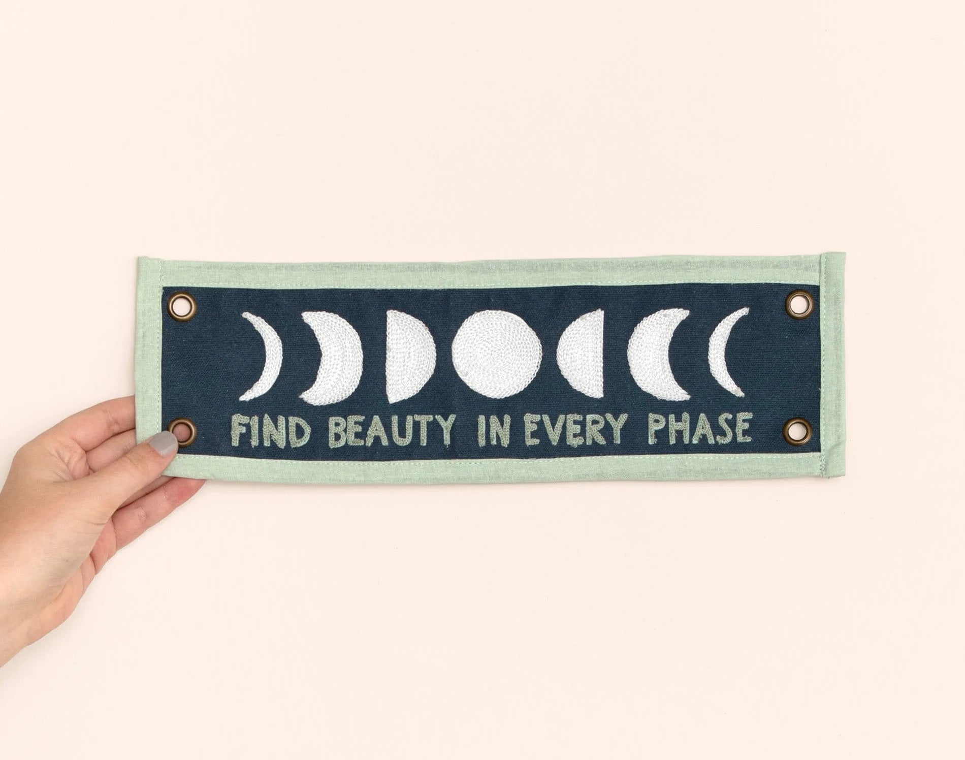 Hand holding the 1canoe2 Find Beauty in Every Phase banner against a peach background