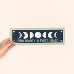 Hand holding the 1canoe2 Find Beauty in Every Phase banner against a peach background