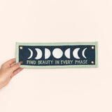 Hand holding the 1canoe2 Find Beauty in Every Phase banner against a peach background