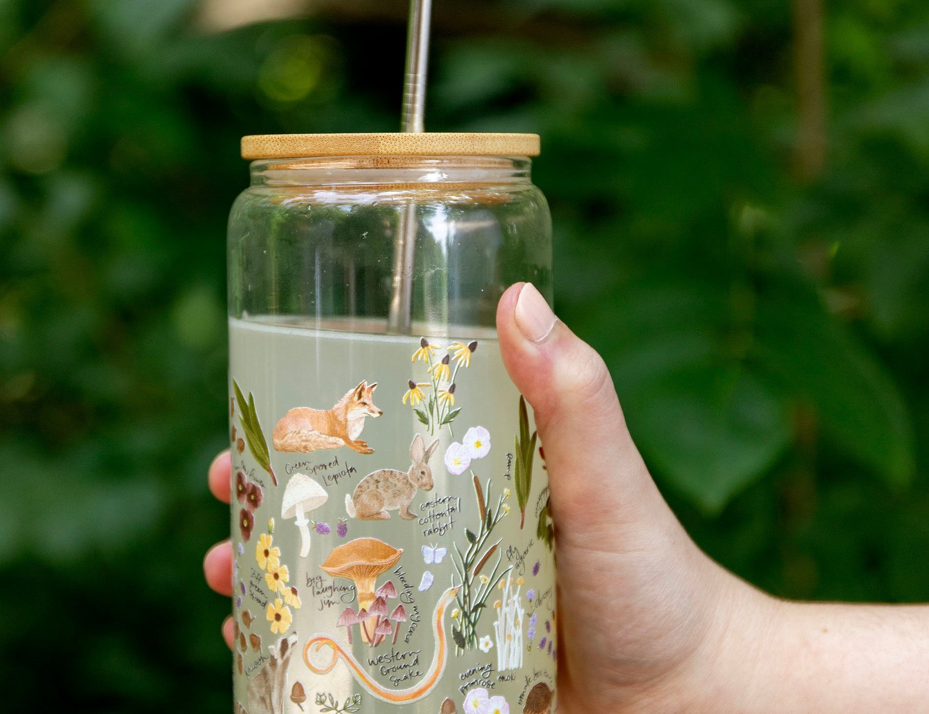 Hand holding the 1canoe2 Flora & Fauna Glass Can with a fox and wildflower print, outdoors