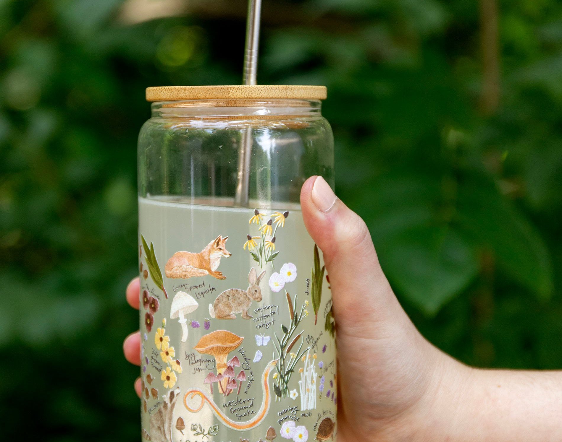 Hand holding the 1canoe2 Flora & Fauna Glass Can with a fox and wildflower print, outdoors