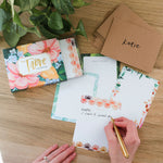 Hands writing on a 1canoe2 Trove Flat Note with kraft envelopes and the floral box