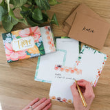 Hands writing on a 1canoe2 Trove Flat Note with kraft envelopes and the floral box