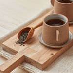 Canyon Clay Cup and Saucer with small handle by Citrine made from exposed reddish-brown earthenware and finished with a thin off-white glaze. Holding tea sitting on cutting board next to loose leaf tea.