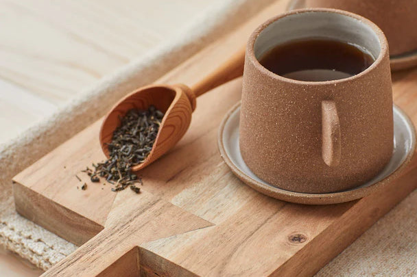 Canyon Clay Cup and Saucer with small handle by Citrine made from exposed reddish-brown earthenware and finished with a thin off-white glaze. Holding tea sitting on cutting board next to loose leaf tea.