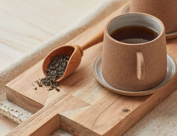 Canyon Clay Cup and Saucer with small handle by Citrine made from exposed reddish-brown earthenware and finished with a thin off-white glaze. Holding tea sitting on cutting board next to loose leaf tea.