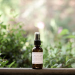 Essential oil and non-toxic Camp Mist Insect Shield by Among the Flowers in amber spray bottle with black lid and white label sitting on wood with green plants in background. 