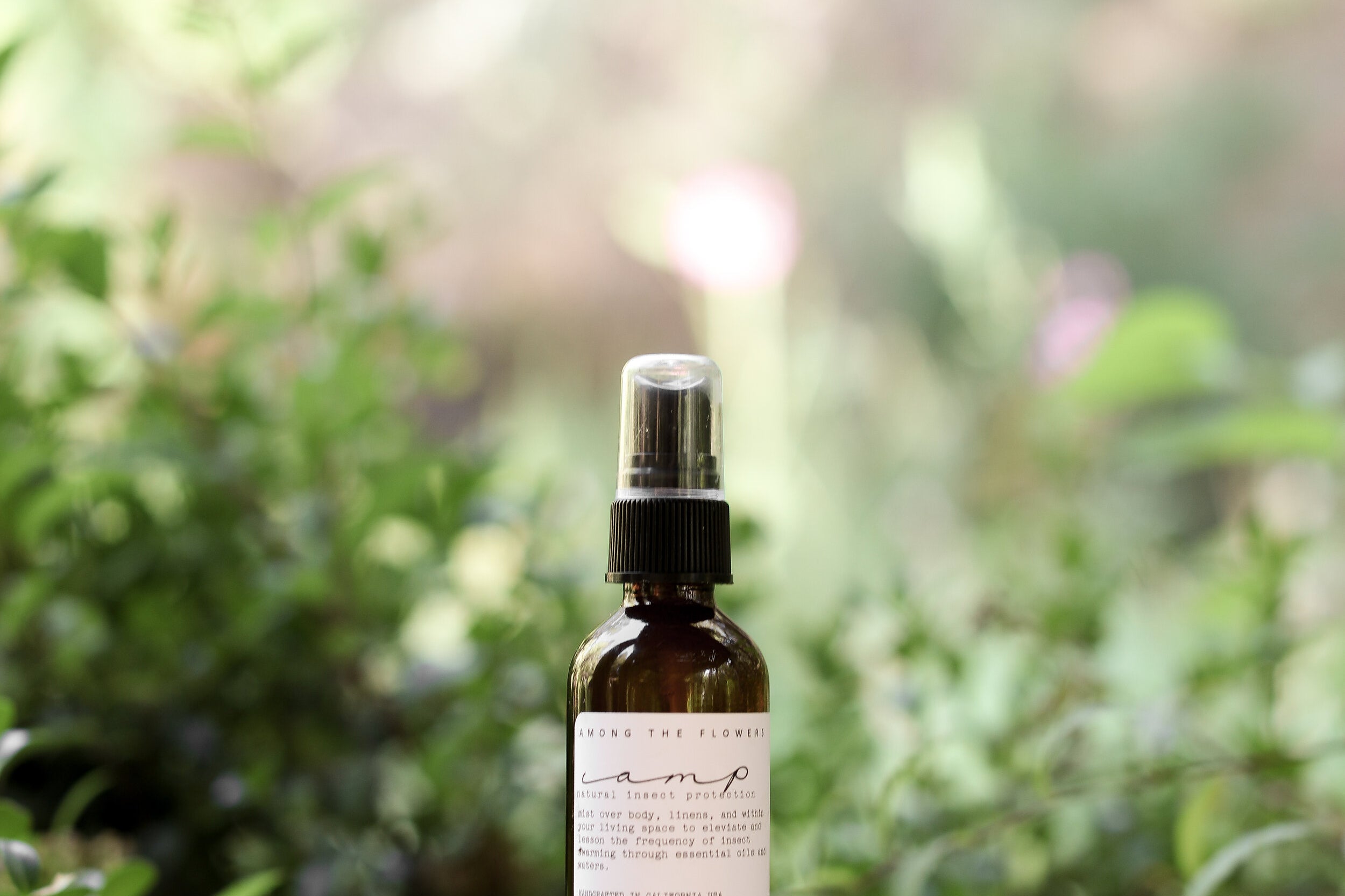 Essential oil and non-toxic Camp Mist Insect Shield by Among the Flowers in amber spray bottle with black lid and white label sitting on wood with green plants in background. 