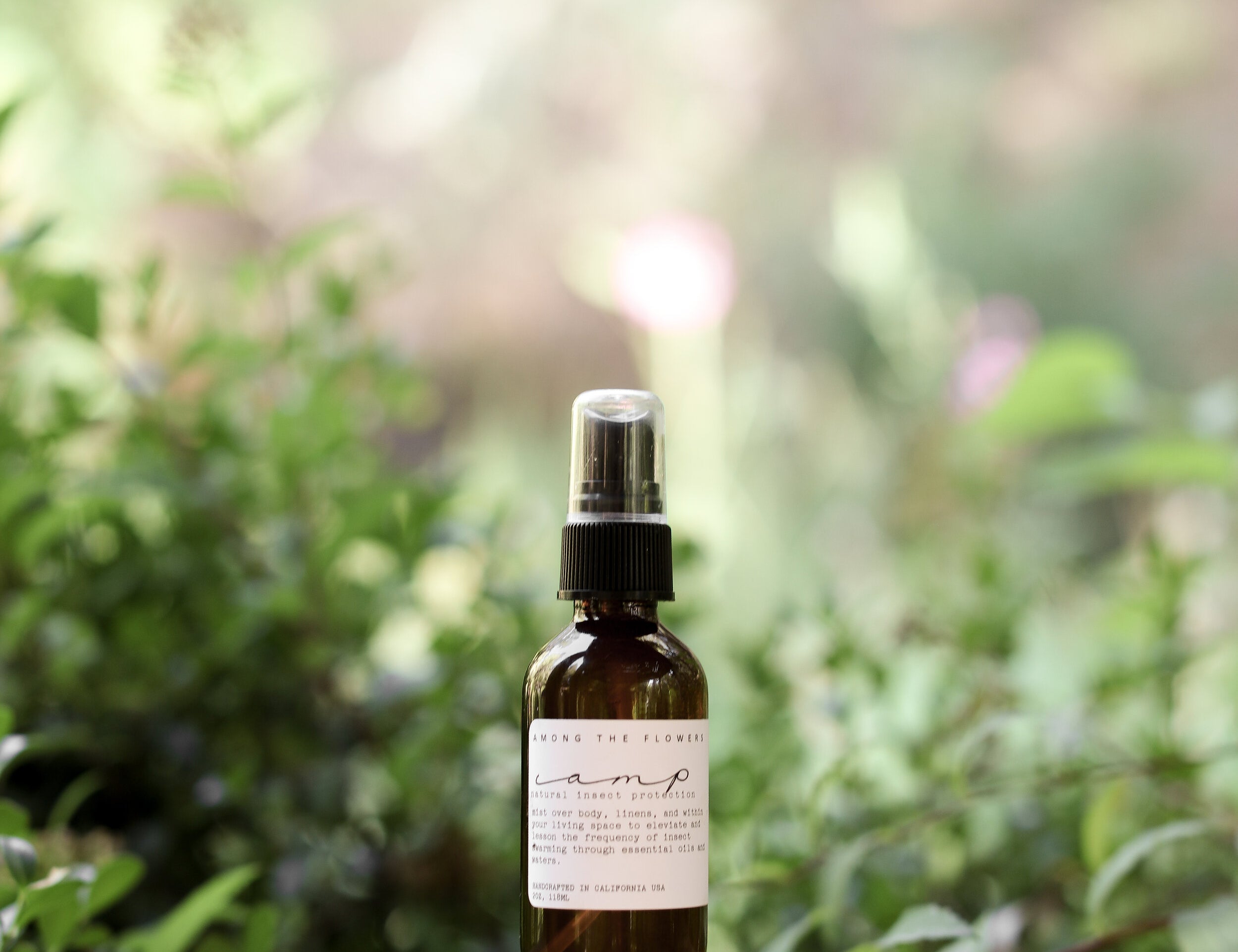 Essential oil and non-toxic Camp Mist Insect Shield by Among the Flowers in amber spray bottle with black lid and white label sitting on wood with green plants in background. 