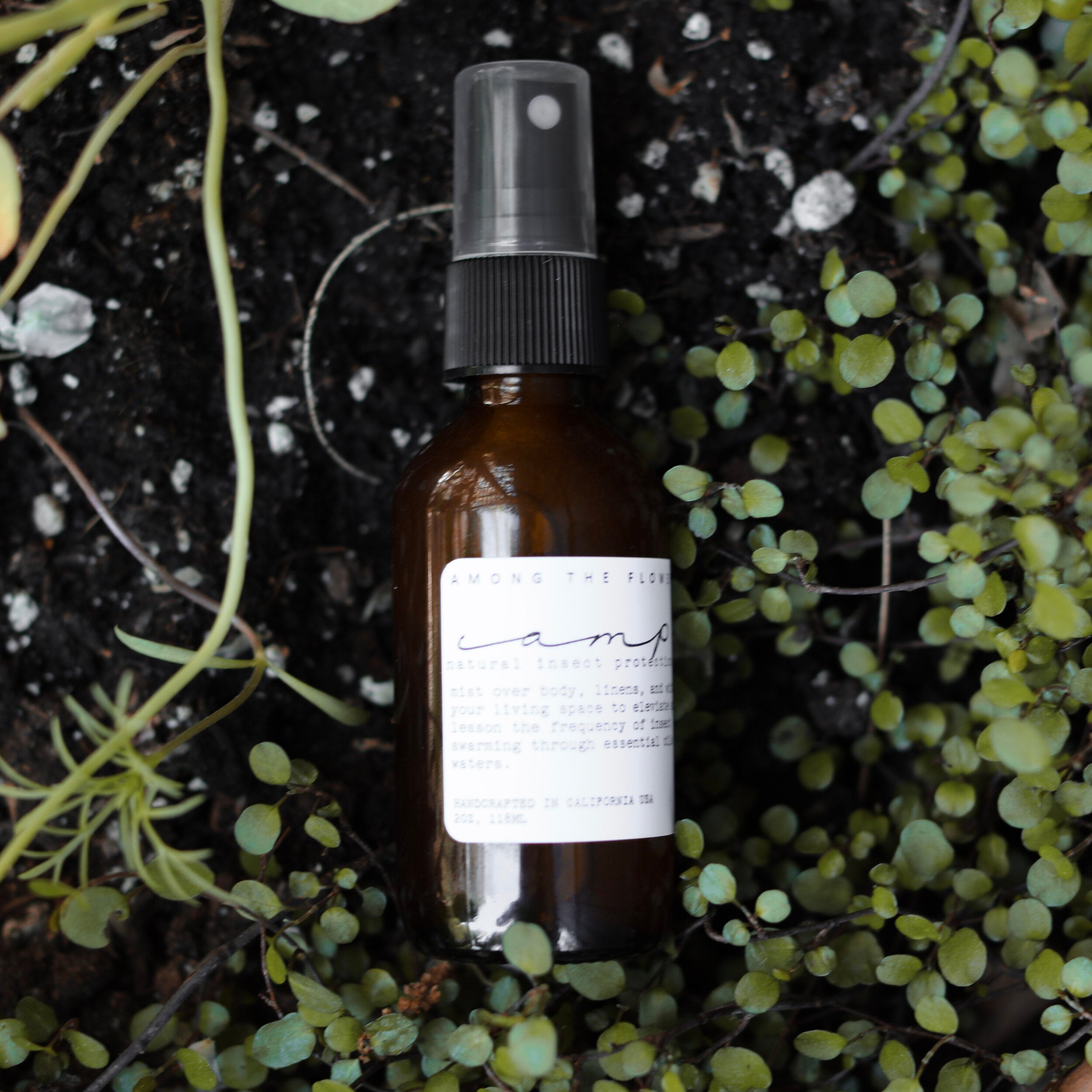 Essential oil and non-toxic Camp Mist Insect Shield by Among the Flowers in amber spray bottle with black lid and white label. Microgreens and soil background.