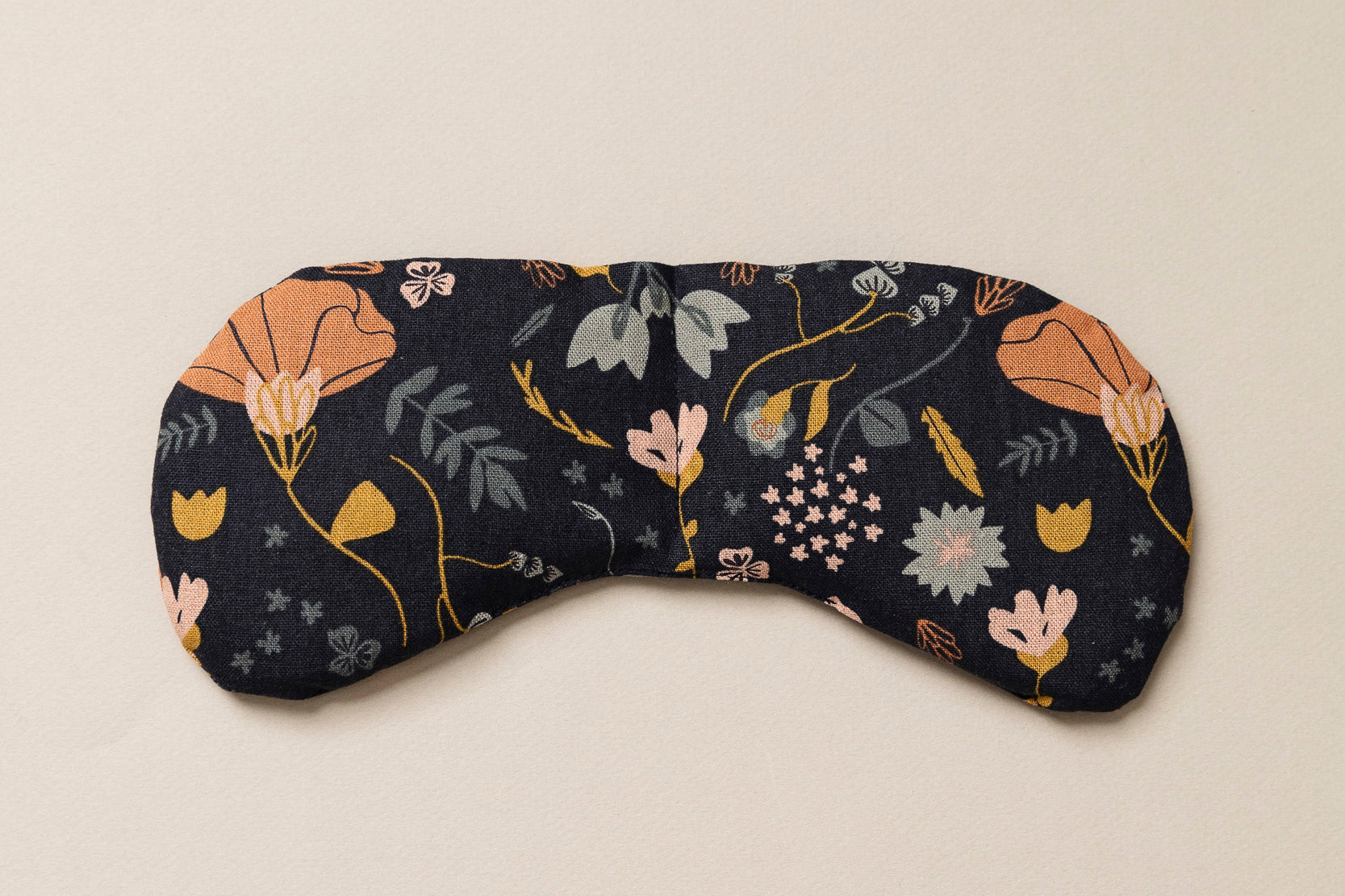Migraine Mask by Slow North. Strap free cotton weighted eye pillow to be used hot or cold to sooth headaches and tired eyes. Navy with multicolor floral design. Tan background.