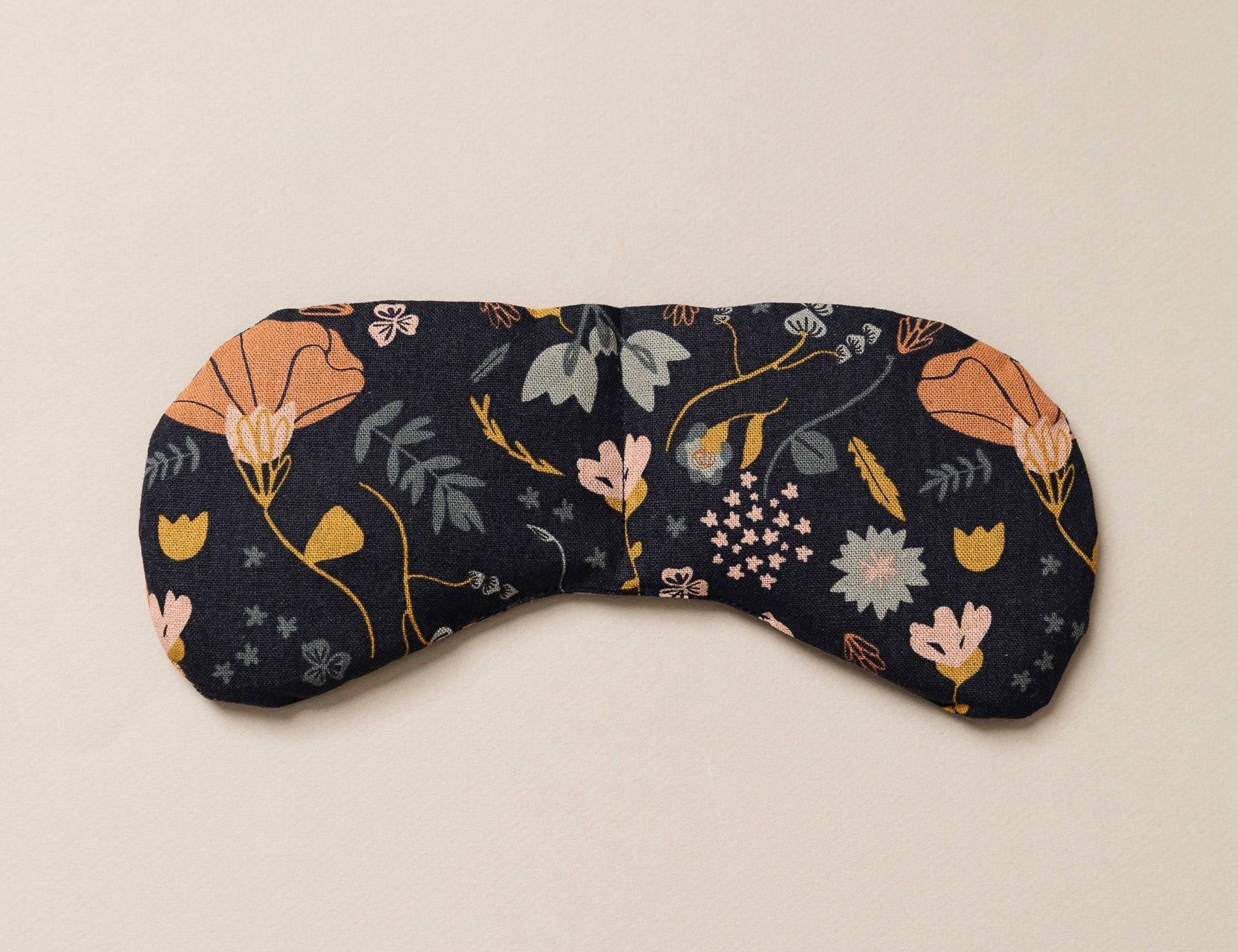 Migraine Mask by Slow North. Strap free cotton weighted eye pillow to be used hot or cold to sooth headaches and tired eyes. Navy with multicolor floral design. Tan background.