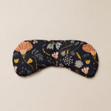 Migraine Mask by Slow North. Strap free cotton weighted eye pillow to be used hot or cold to sooth headaches and tired eyes. Navy with multicolor floral design. Tan background.