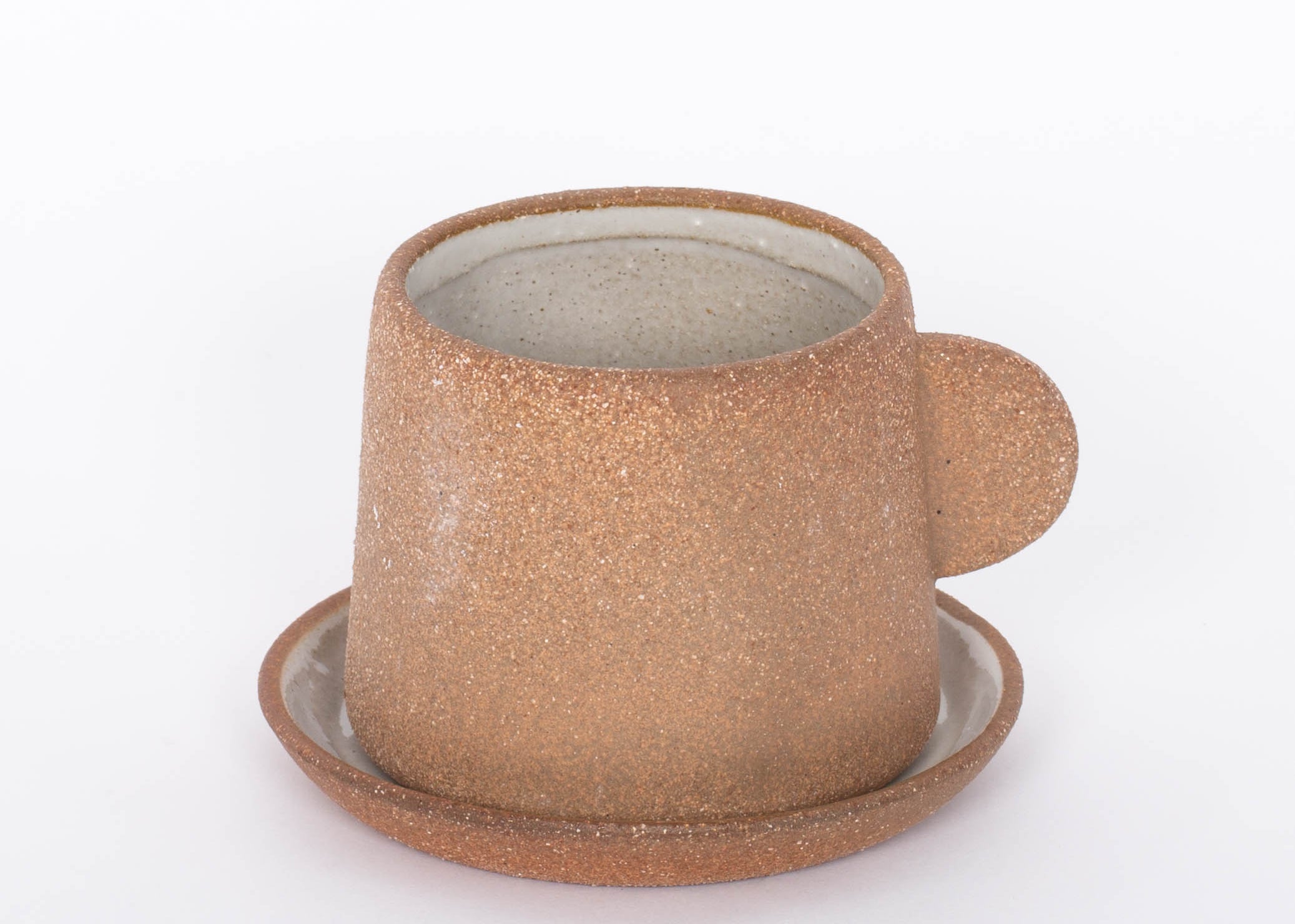 Canyon Clay Cup and Saucer with small handle by Citrine made from exposed reddish-brown earthenware and finished with a thin off-white glaze. 