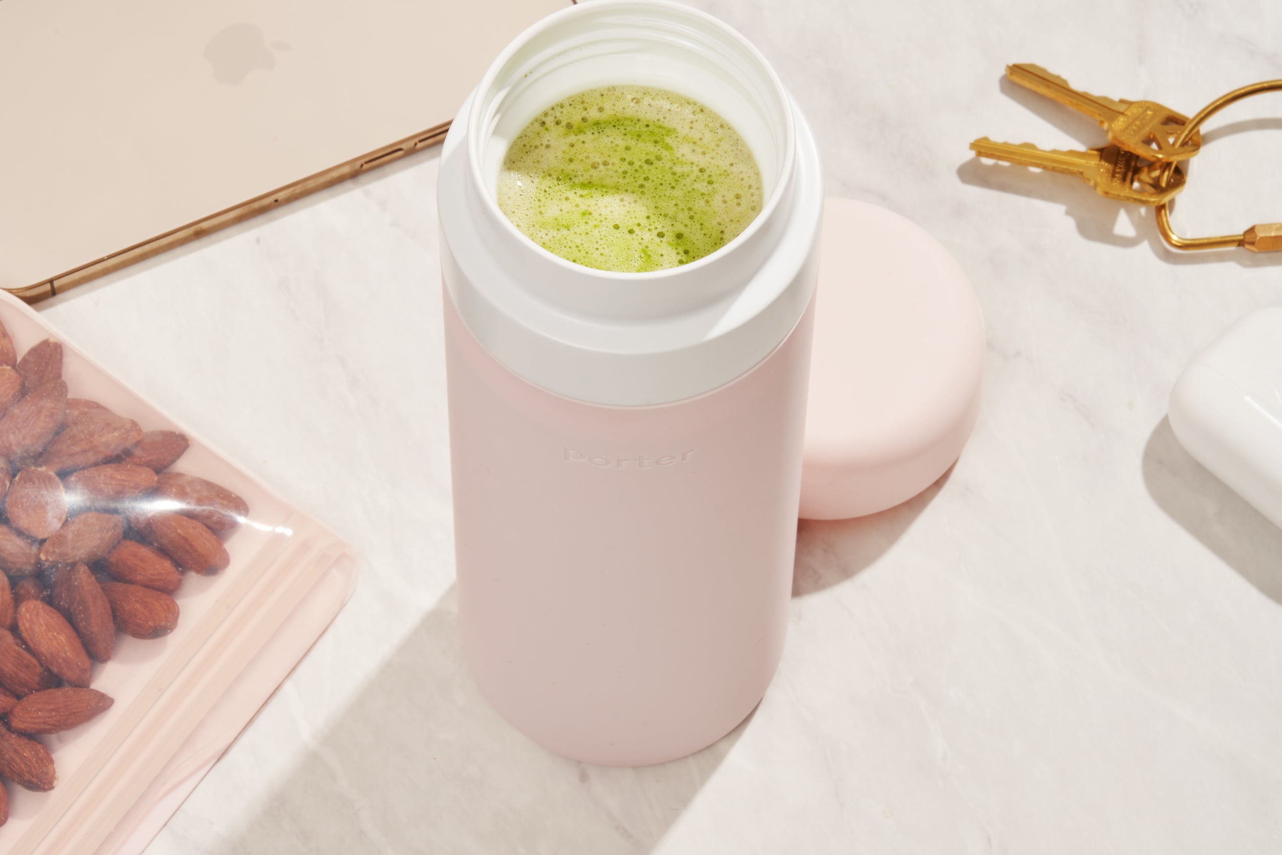 Blush wide mouth ceramic Porter waterbottle with green matcha latte inside. Phone, keys, and bag of almonds on counter. 