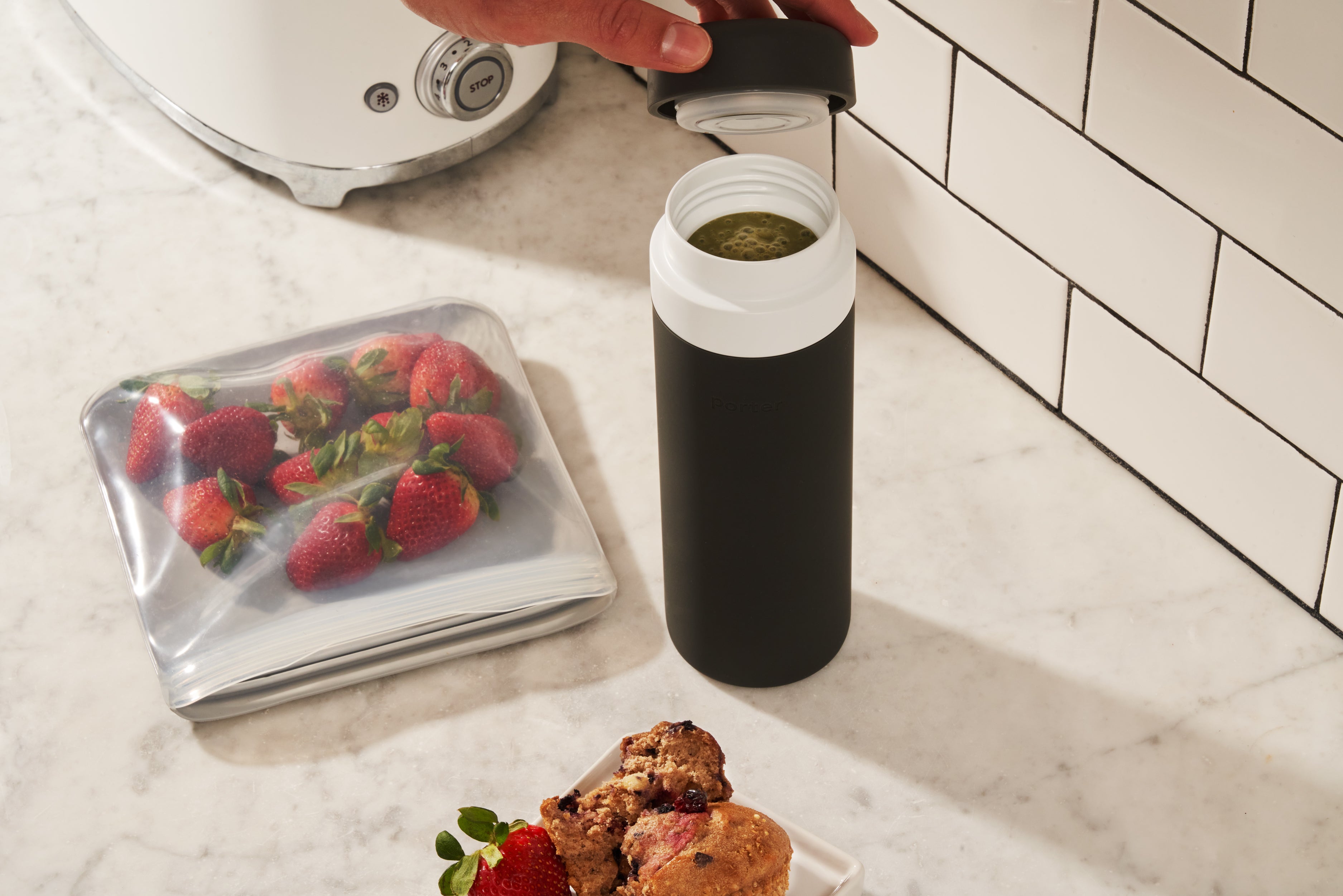 Person putting black silicone leakproof lid on Porter Insulated Charcoal Waterbottle with white top, next to kitchen counter and strawberries. 