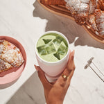 Woman's hand holding blush Porter ceramic wide mouth waterbottle with iced matcha latter and powered croissants on a platter.