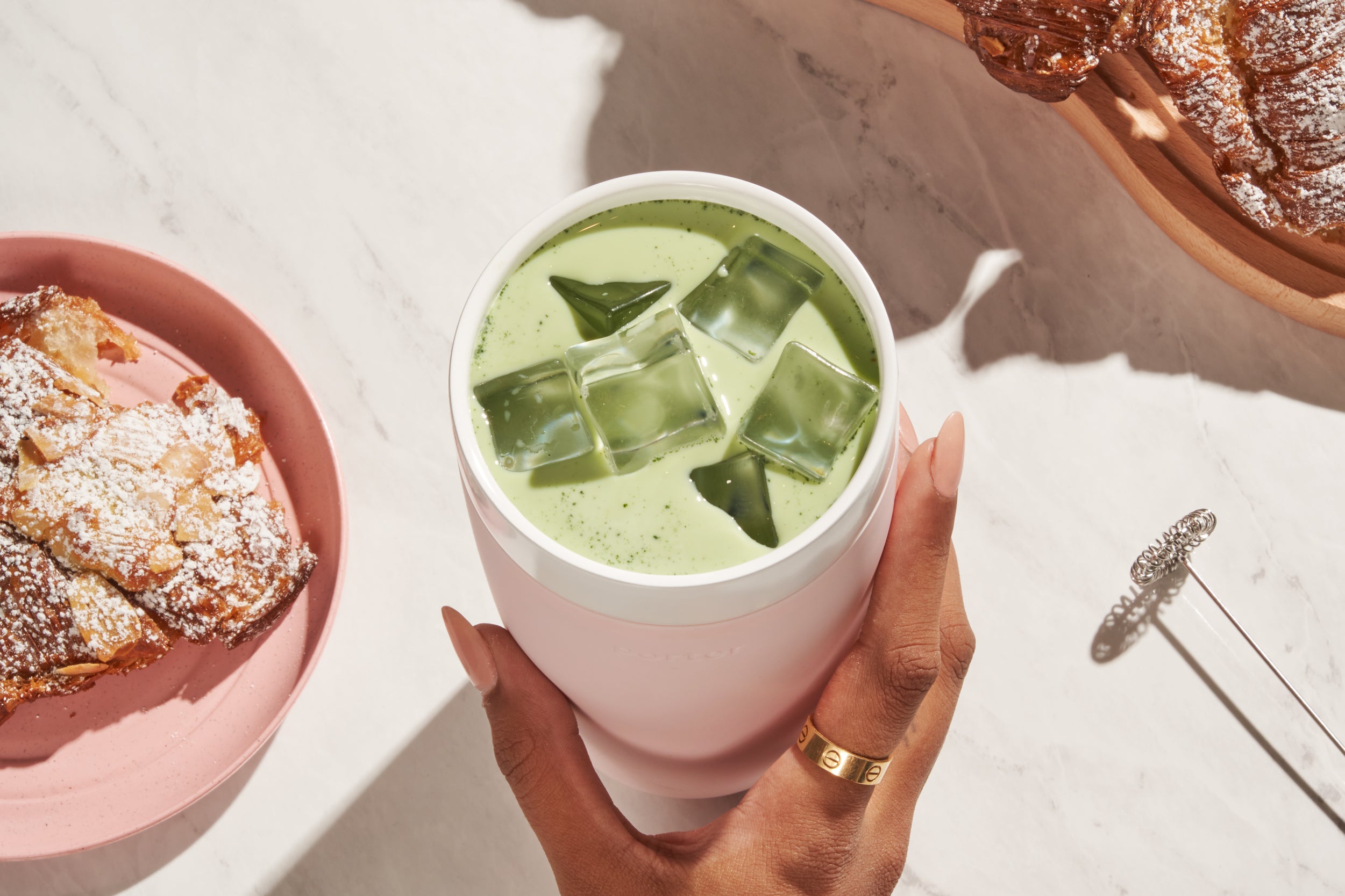 Woman's hand holding blush Porter ceramic wide mouth waterbottle with iced matcha latter and powered croissants on a platter.