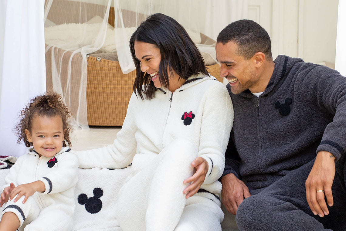 Family of three sitting on a wooden deck with a white blanket featuring black mouse ears and tails.