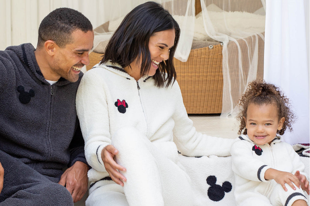 A family of three wearing matching Mickey Mouse-themed fleece pajamas, sitting together on a white blanket in a brightly lit room. The father wears a dark gray set, while the mother and young child wear white sets, both featuring black Mickey head silhouettes and a small red bow-tie accent.