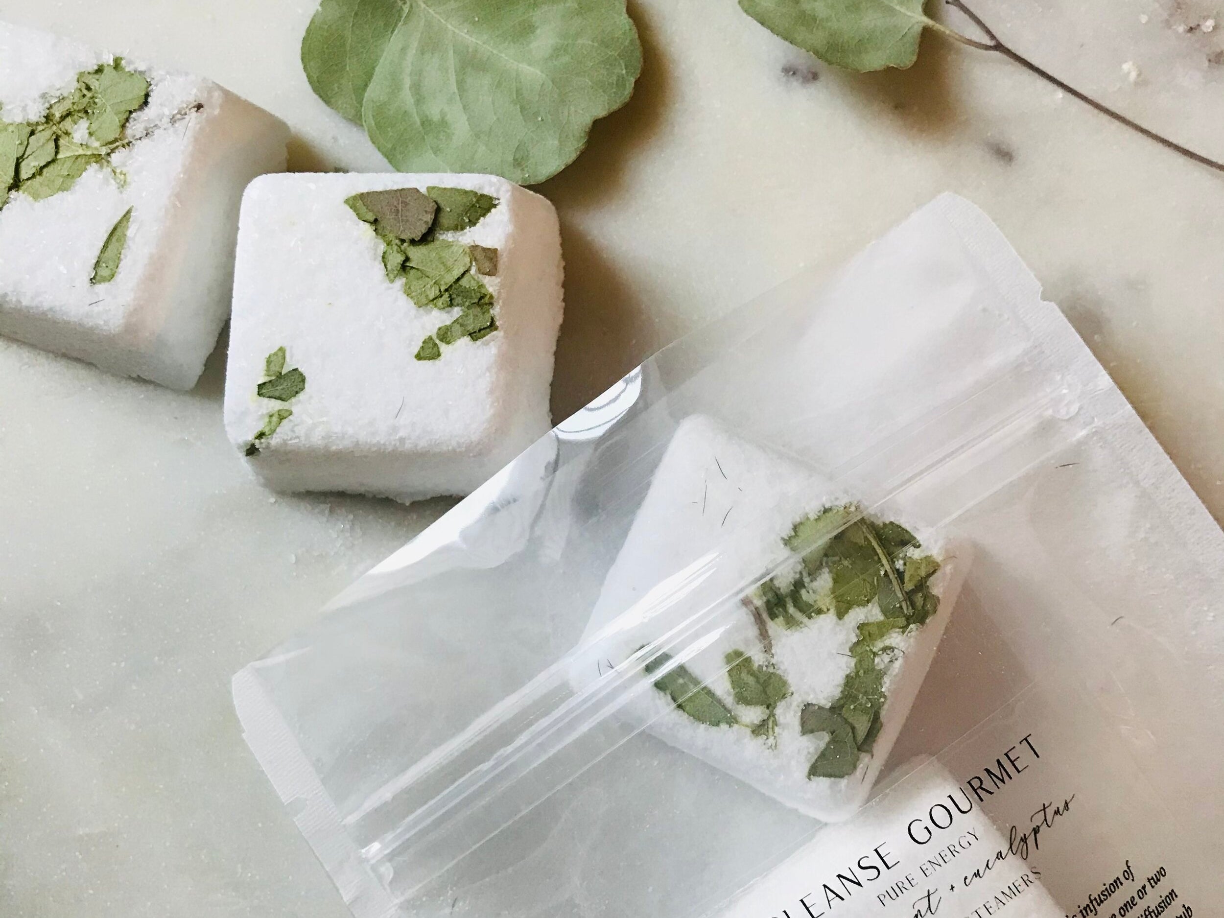 Clear pack of five white and green Pure Energy Eucalyptus + Peppermint Shower Steamers with essential oils by Cleanse Gourmet on counter with eucalyptus leaves. 