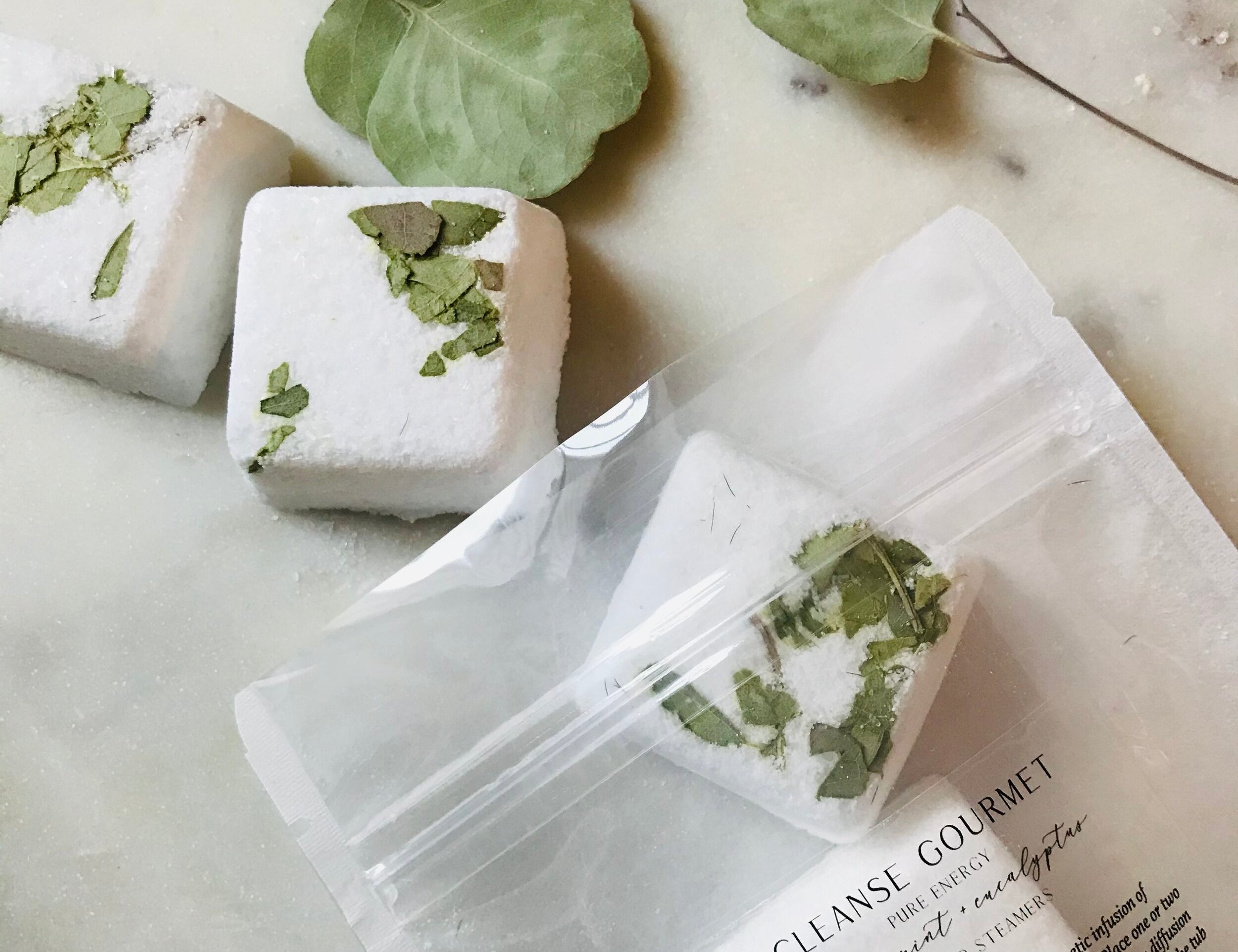 Clear pack of five white and green Pure Energy Eucalyptus + Peppermint Shower Steamers with essential oils by Cleanse Gourmet on counter with eucalyptus leaves. 