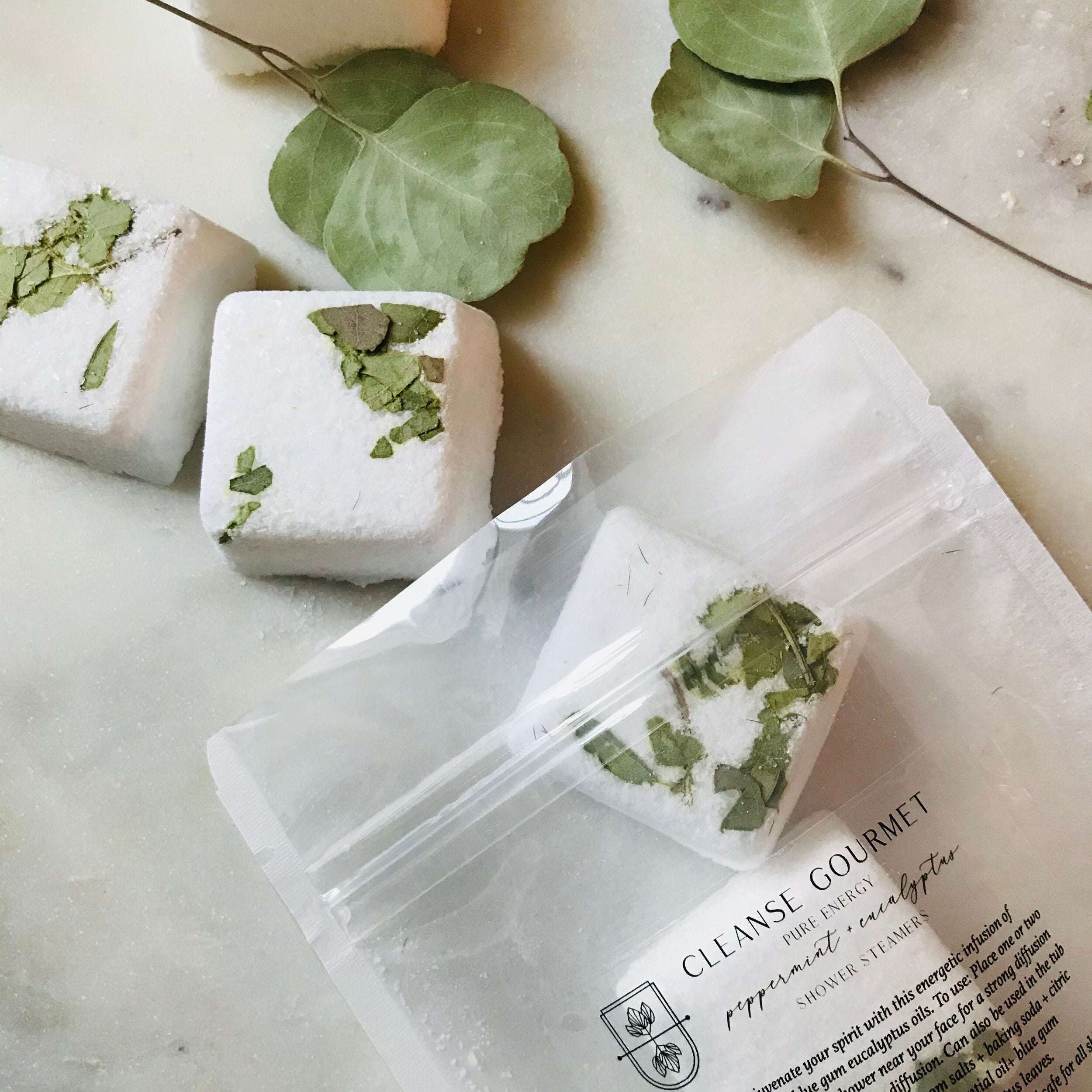 Clear pack of five white and green Pure Energy Eucalyptus + Peppermint Shower Steamers with essential oils by Cleanse Gourmet on counter with eucalyptus leaves. 