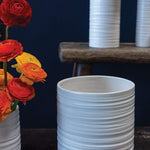 Modern and short striated ceramic Everest Pot in a matte white glaze. Several sizes with red and orange bouquet. Dark navy background. 