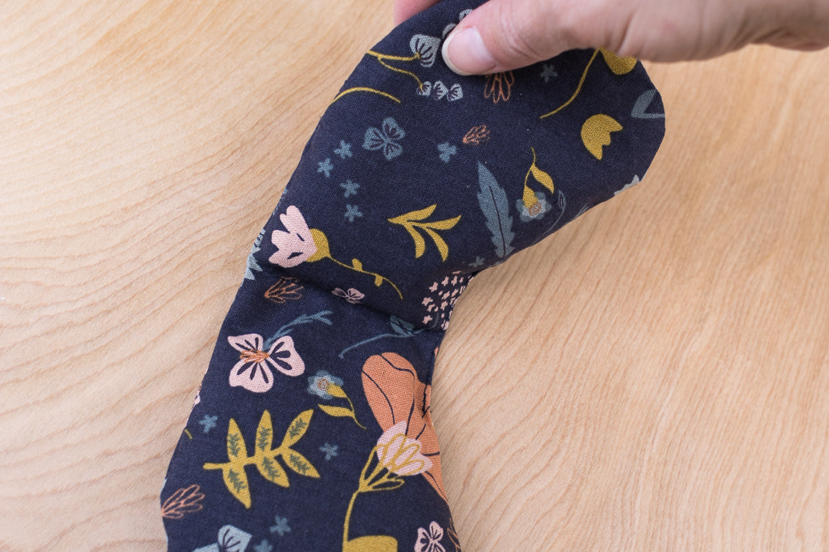 Hand holding Slow North migraine mask over wooden background. Eye pillow is navy with floral design. 