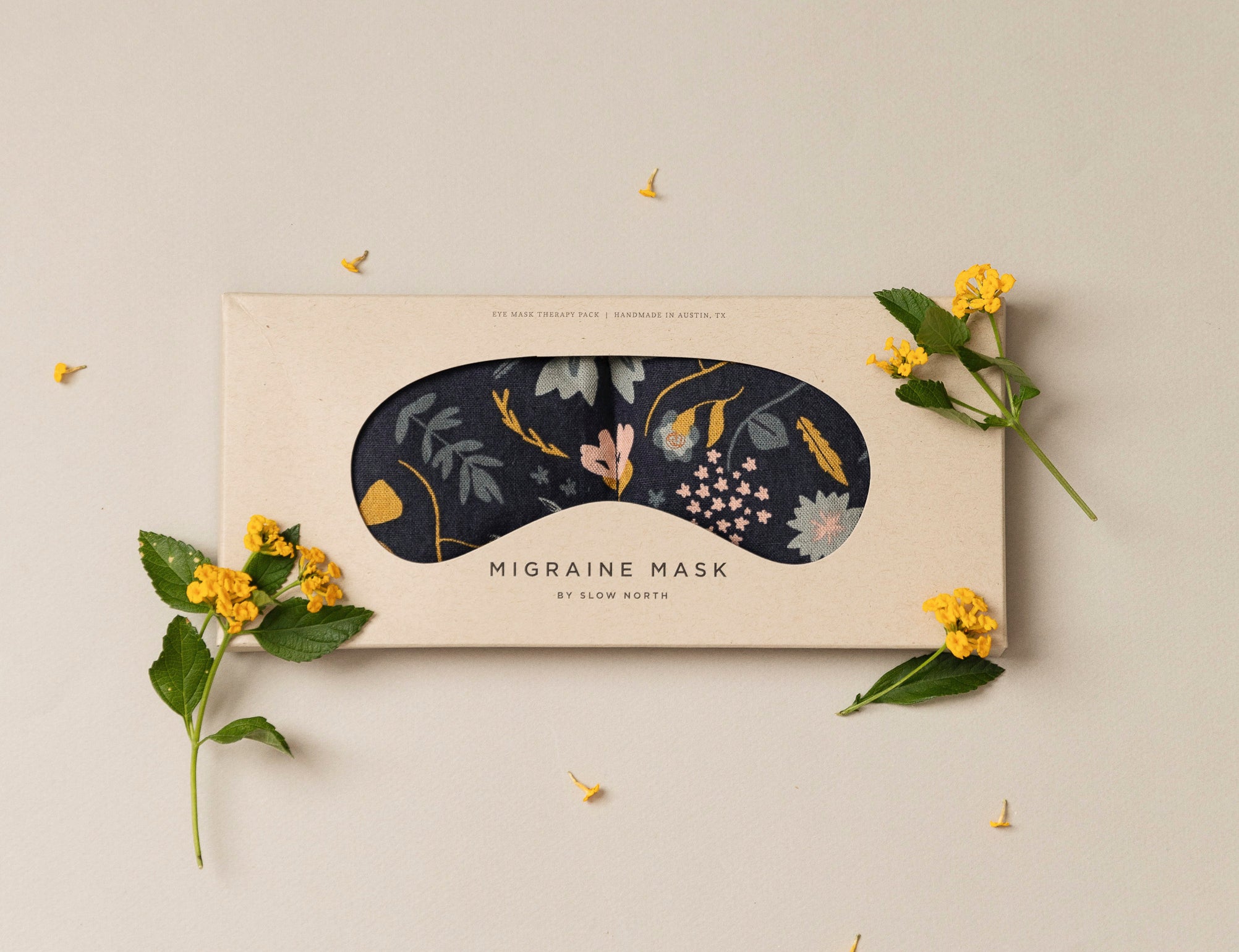 Migraine Mask by Slow North. Strap free cotton weighted eye pillow to be used hot or cold to sooth headaches and tired eyes. Navy and floral print. Inside box surrounded by yellow flowers. 