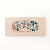 Migraine Mask by Slow North. Strap free cotton weighted eye pillow to be used hot or cold to sooth headaches and tired eyes. Tan and pink and green floral pattern. Inside box. White background.