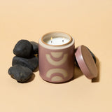 Paddywax blush colored Mandarin Mango Candle. Lit candle with lid off and surrounded by black rocks.