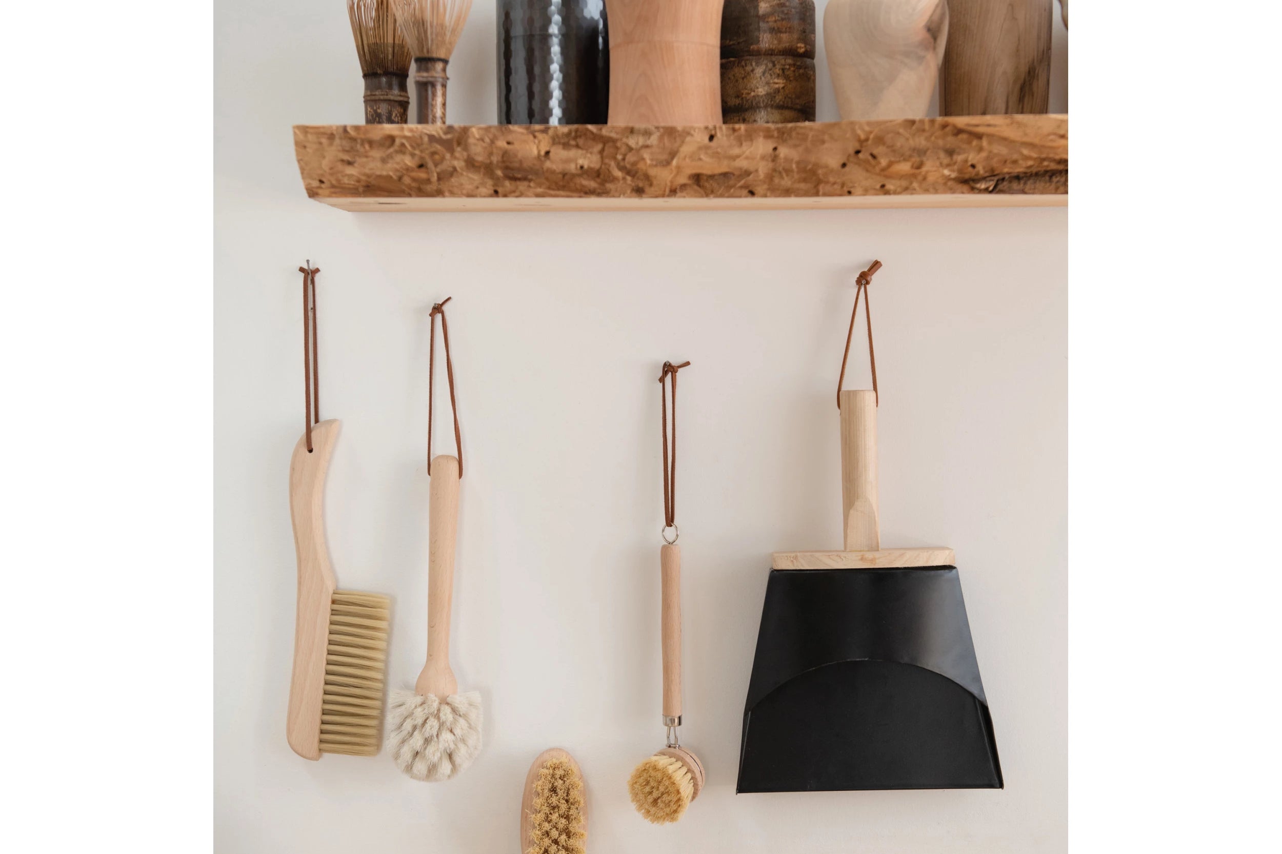 Beech Wood Flat Dish Brush with Leather Strap and silver accents hanging with dustpan and other natural cleaning brushed in neutral washing room.