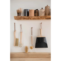 Beech Wood Flat Dish Brush with Leather Strap and silver accents hanging with dustpan and other natural cleaning brushed in neutral washing room.