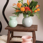 Glenna Vases handcrafted with powdery glaze. Three shown in light green, light pink, and light blue holding an orange bouquet. Set in home on wooden stool and fur rug. 