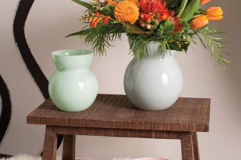 Glenna Vases handcrafted with powdery glaze. Three shown in light green, light pink, and light blue holding an orange bouquet. Set in home on wooden stool and fur rug. 