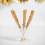 Three Glittering Gold Rock Candy sticks standing in glass with gold streamers in background. 