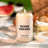 Lit Gone Hiking Candle by Homesick. Glass jar with white candle and bold "GONE HIKING" label. Set on checked table cover. 
