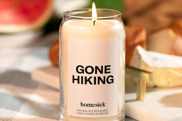 Lit Gone Hiking Candle by Homesick. Glass jar with white candle and bold "GONE HIKING" label. Set on checked table cover. 
