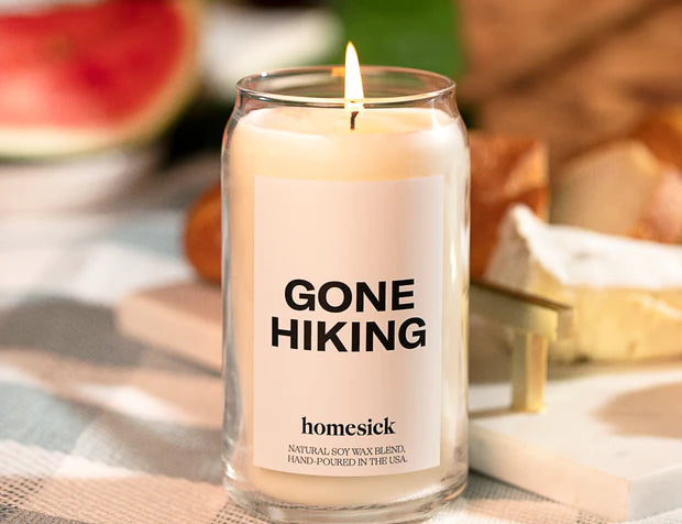 Lit Gone Hiking Candle by Homesick. Glass jar with white candle and bold "GONE HIKING" label. Set on checked table cover. 