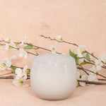 16oz Heure du Thé Ballon candle by Alixx in white. Lily of the valley, water cedar, and musk fragrance. White flowers and pink background behind.